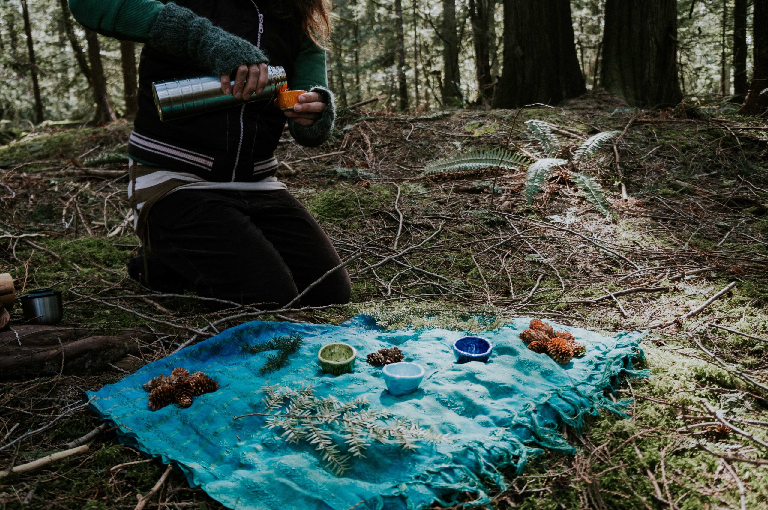 A woman pours liquid from a thermos into a small cup while kneeling on the forest floor. In front of her is a blanket, upon which sit various objects such as pinecones, small tree branches, and small ceramic bowls.