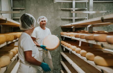 Two workers in a cheese factory proudly display a large wheel of cheese they are holding together.