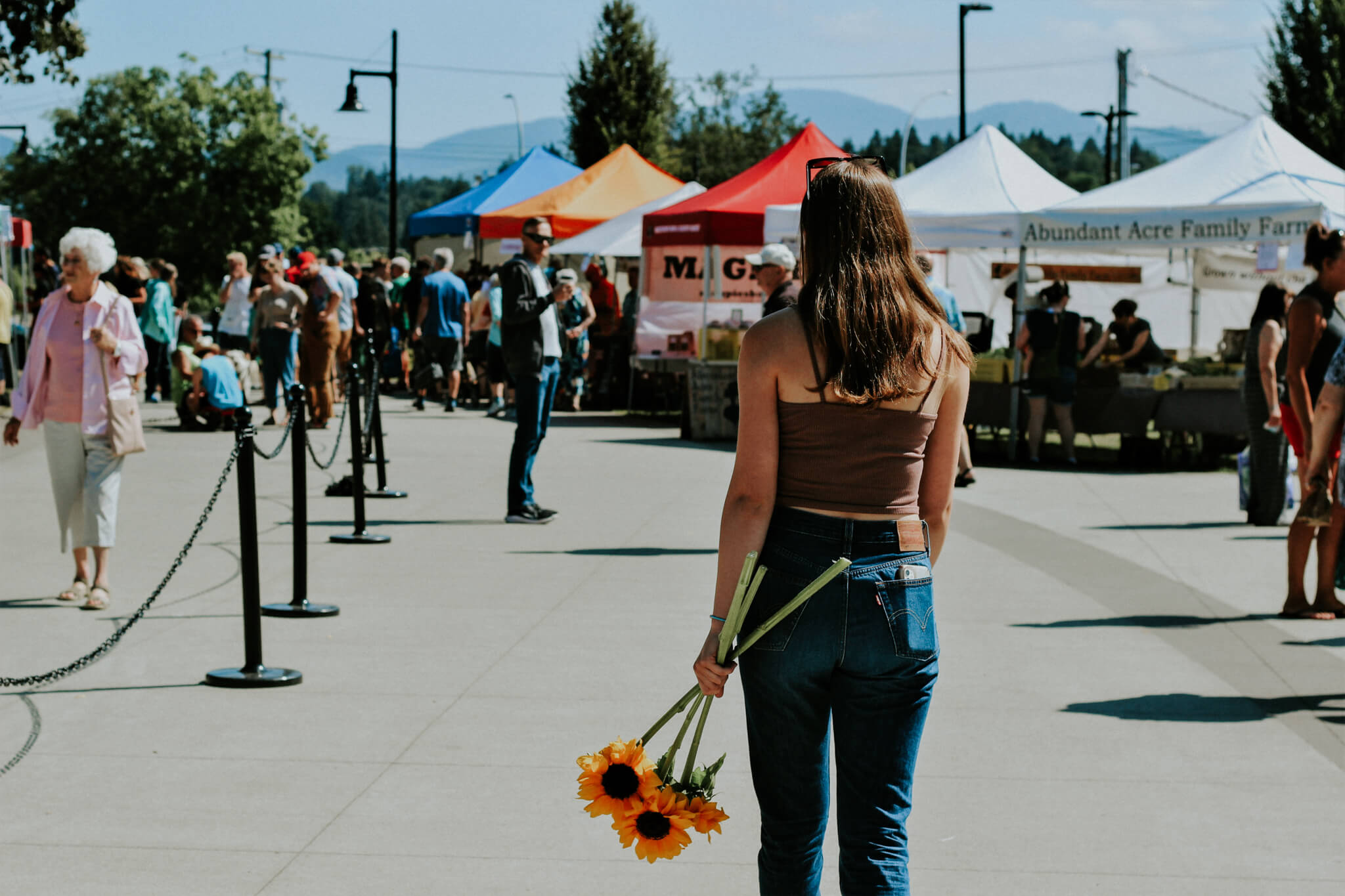 A woman walks through a farmers market on a bright sunny day, carrying a bouquet of sunflowers.