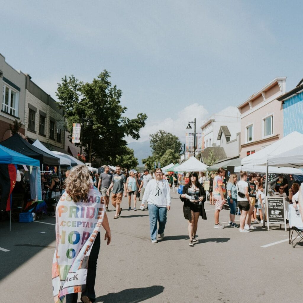 A person wearing a pride flag around their neck walks down the middle of a street, with various tents on the sides.