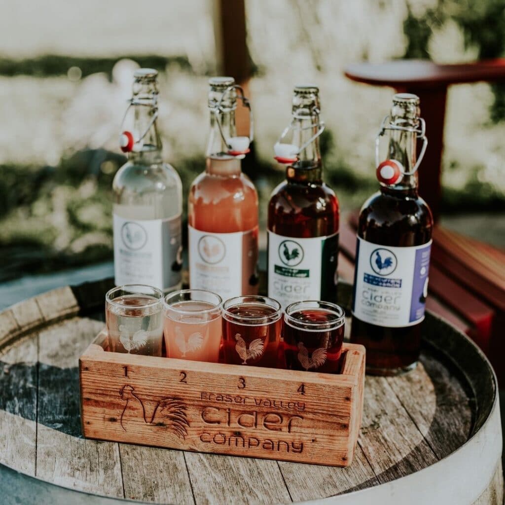 A flight of ciders in a wooden box, with the bottles of each corresponding cider behind them