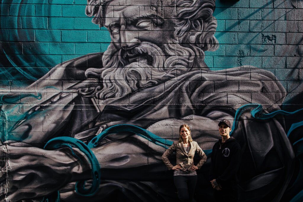 Two people stand in front of an outdoor mural.