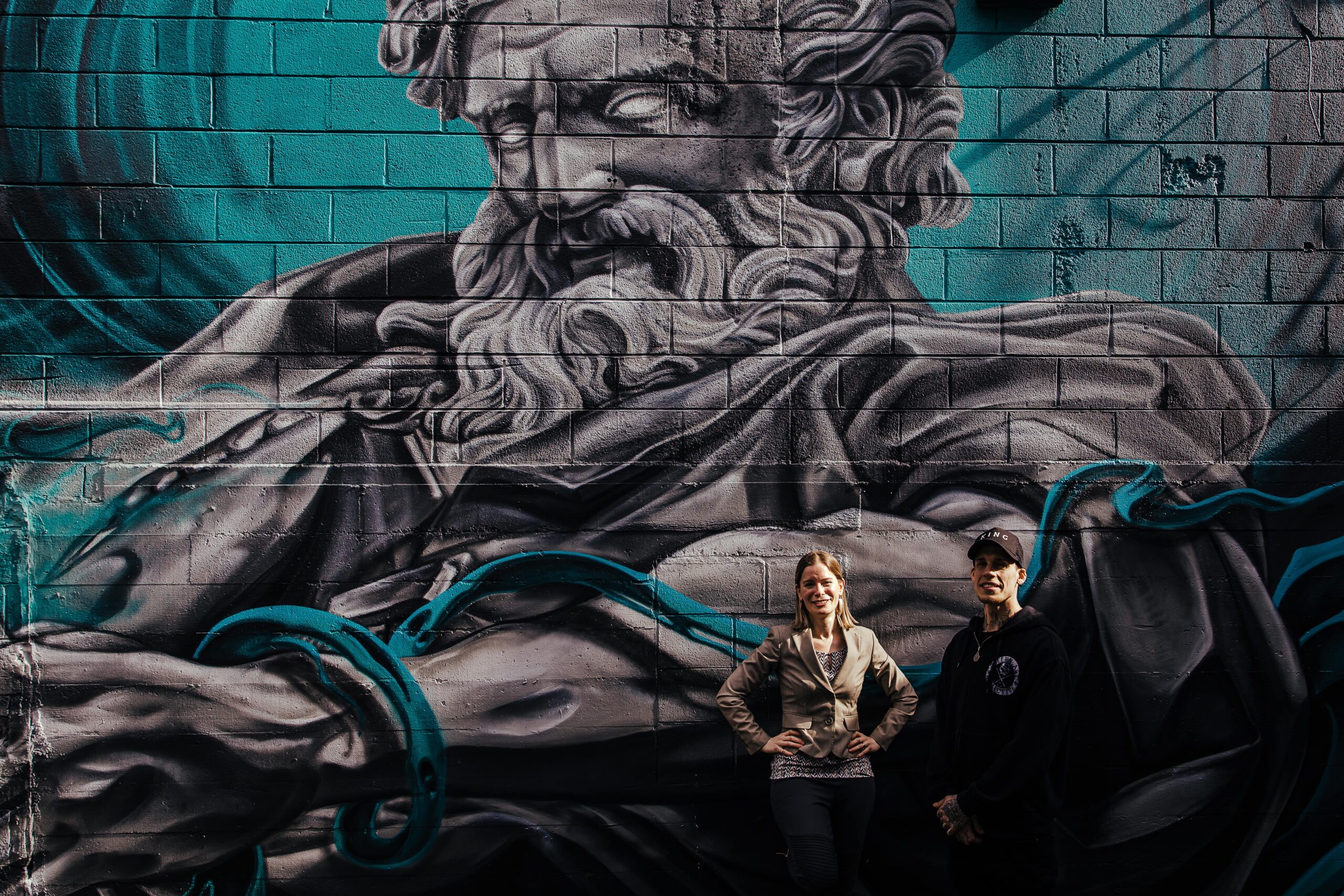 Two people stand in front of an outdoor mural.