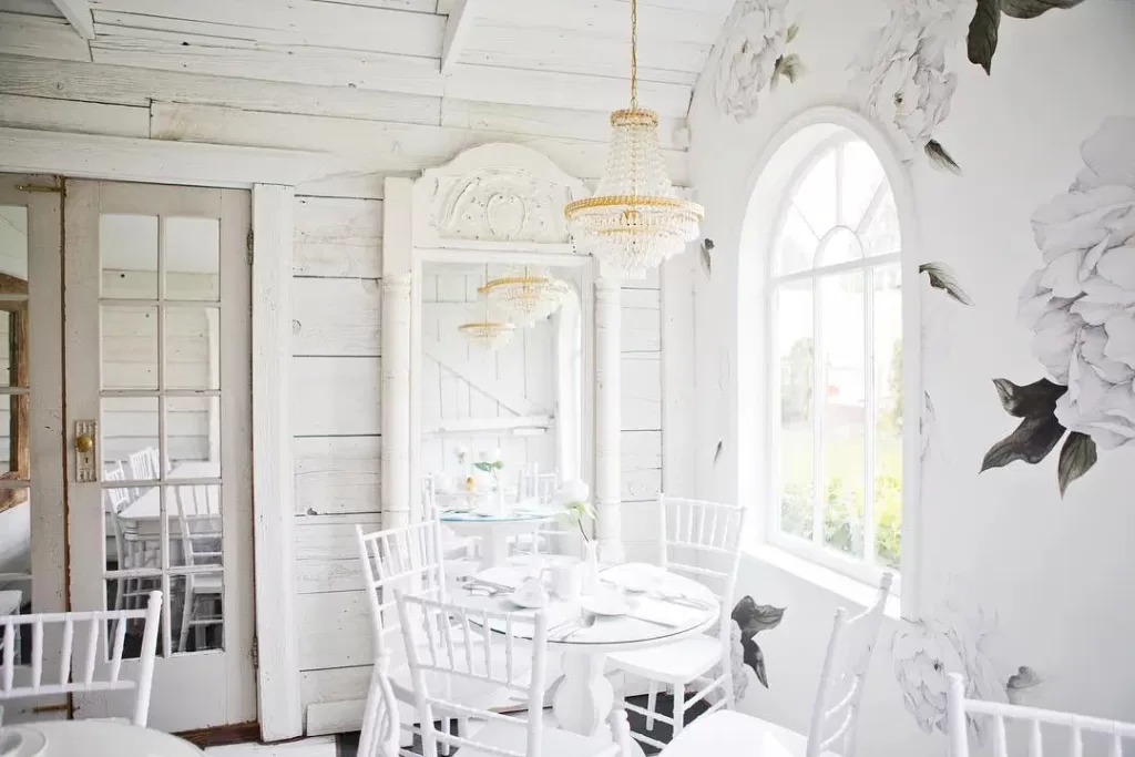 The dining area of Little White House & Co, which features all-white wooden chairs and tables, white walls and furnishings, and a glass chandelier.