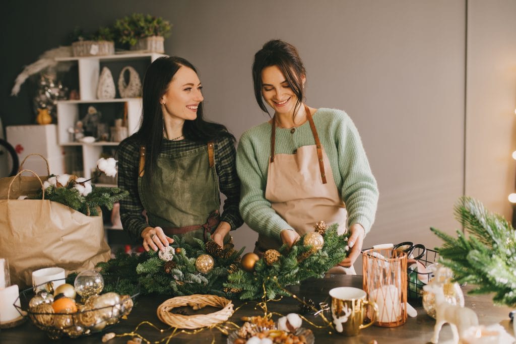 Two Millennial Women Making Christmas Wreath Using Pine Branches And Festive Decorations