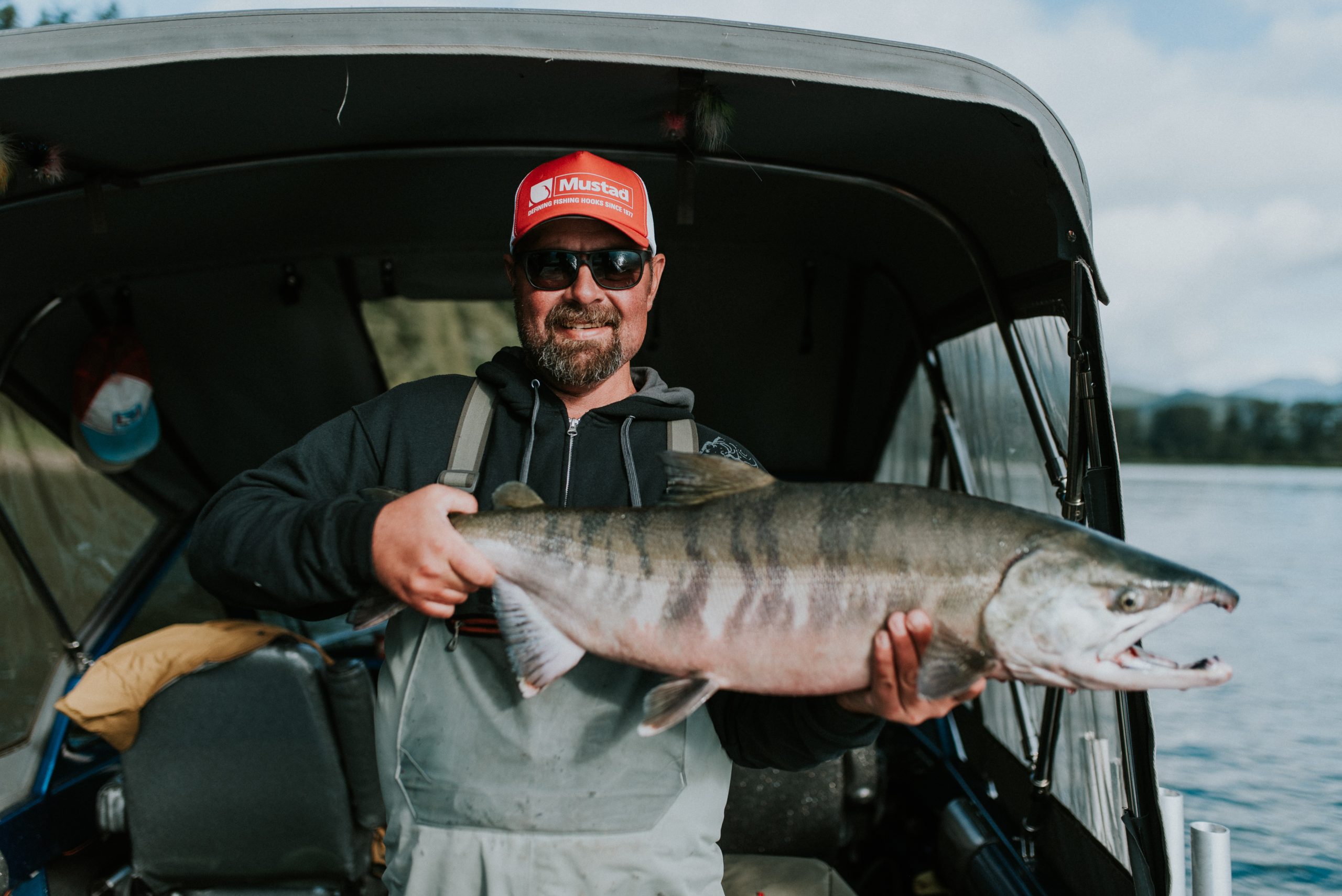 A man, wearing a red hat, sunglasses, and overalls proudly and happily faces the camera as he holds a large fish in his hands