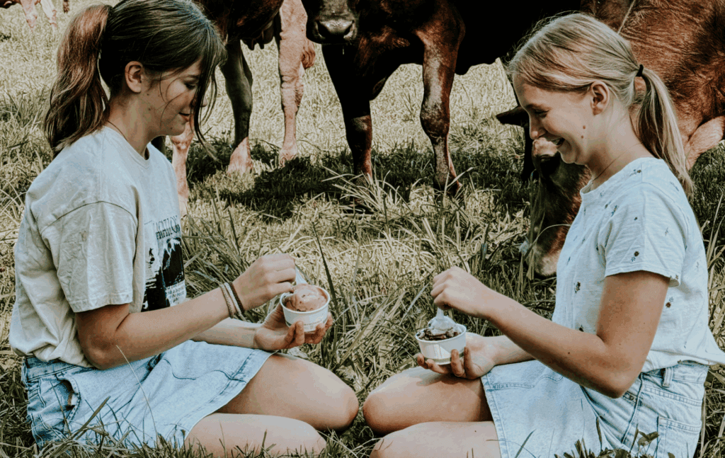 Two girls sitting and eating ice cream on the grass with cows behind them