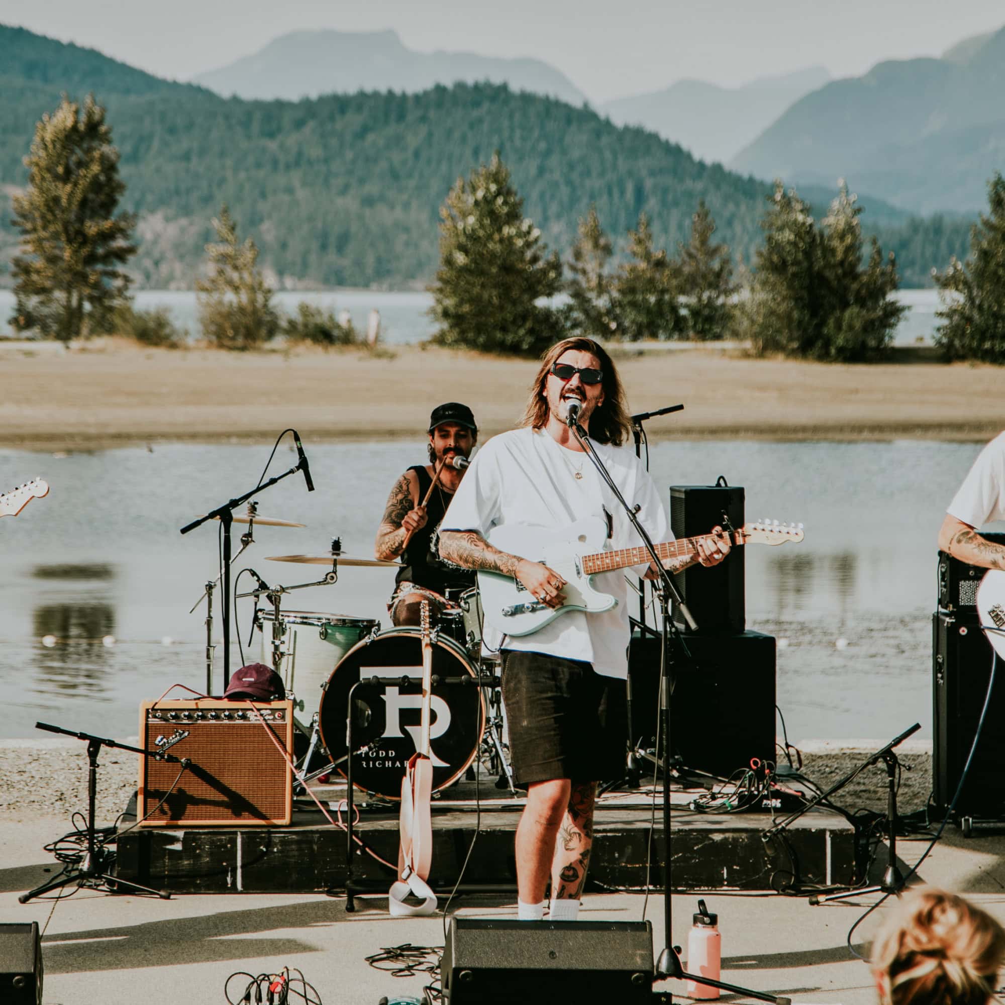 a man playing a guitar sings into a mic, in an outdoor setting, mountains and a lake in the background
