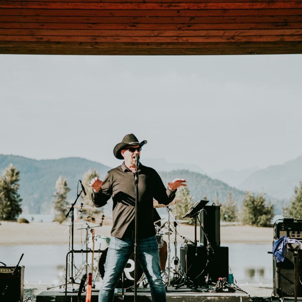 A man with a cowboy hat sings into a mic with a lake and mountains in the background