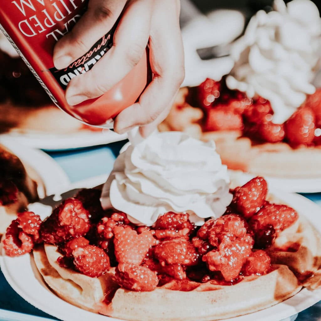 A person pouring whipped cream on a strawberry waffle.