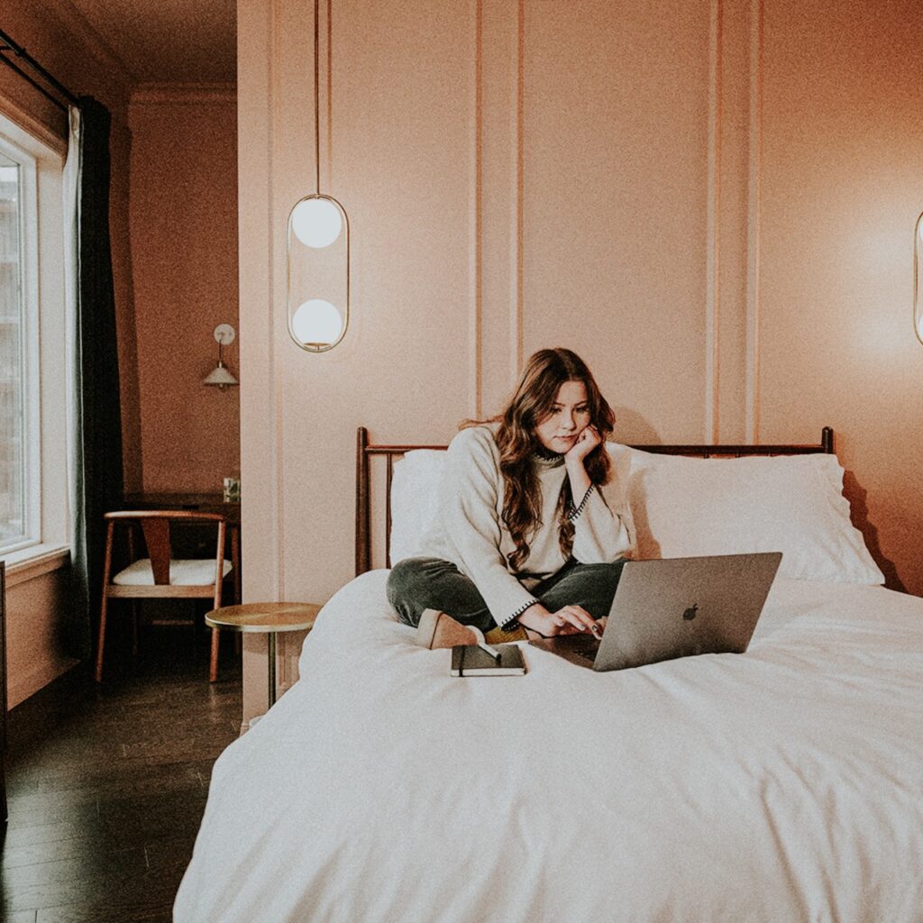 A woman sits on a bed looking at her laptop
