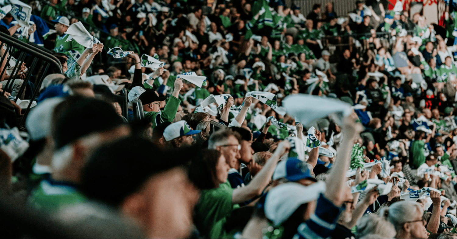 Fans wearing team jerseys cheer from the stands at a hockey game inside a packed arena.
