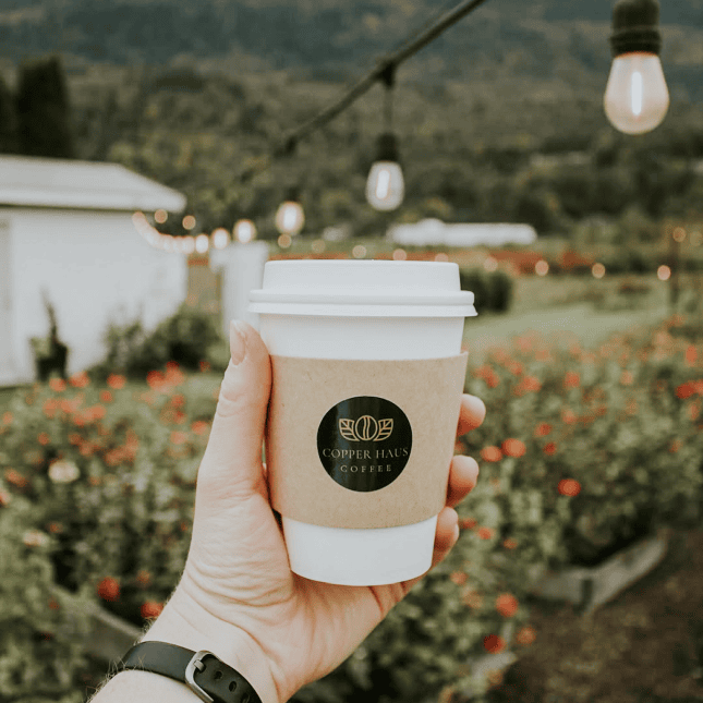 Hand holding a takeaway coffee cup outdoors with flowers and mountains in the background.