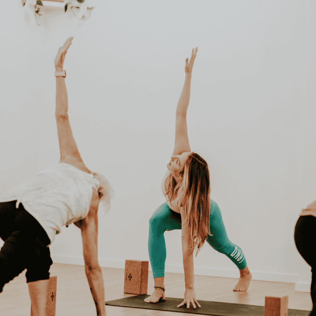 Two people practicing yoga, holding a side stretch pose in a bright studio.