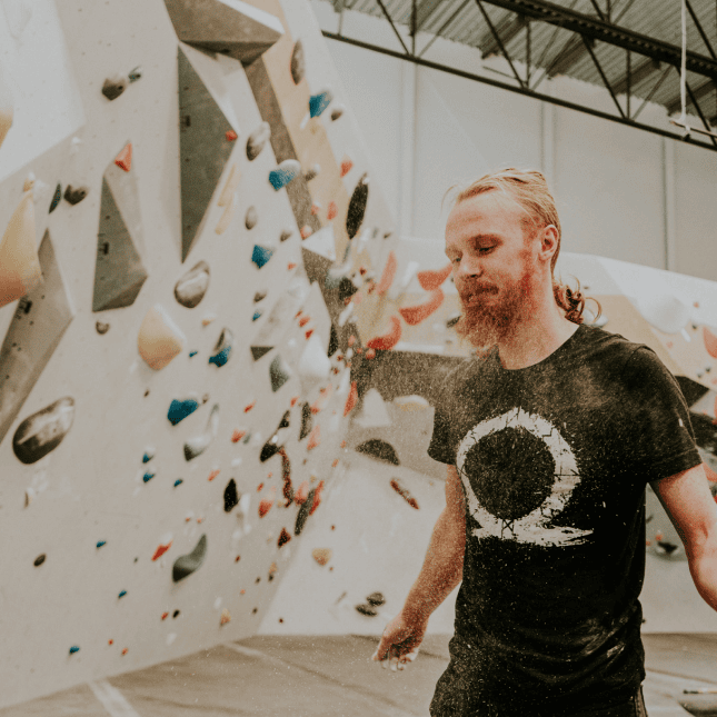 A person smiling while standing beneath an indoor climbing wall.