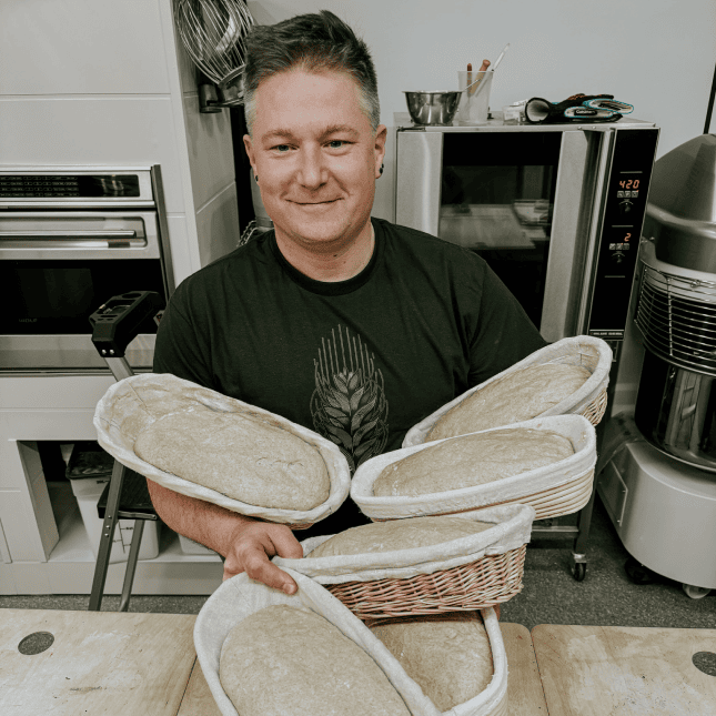 Chilliwack Baker holding trays of freshly baked bread inside a commercial kitchen.