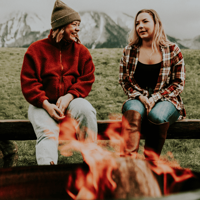 Two people sitting by an outdoor fire pit, bundled in warm clothing, with a rustic lodge setting behind them.