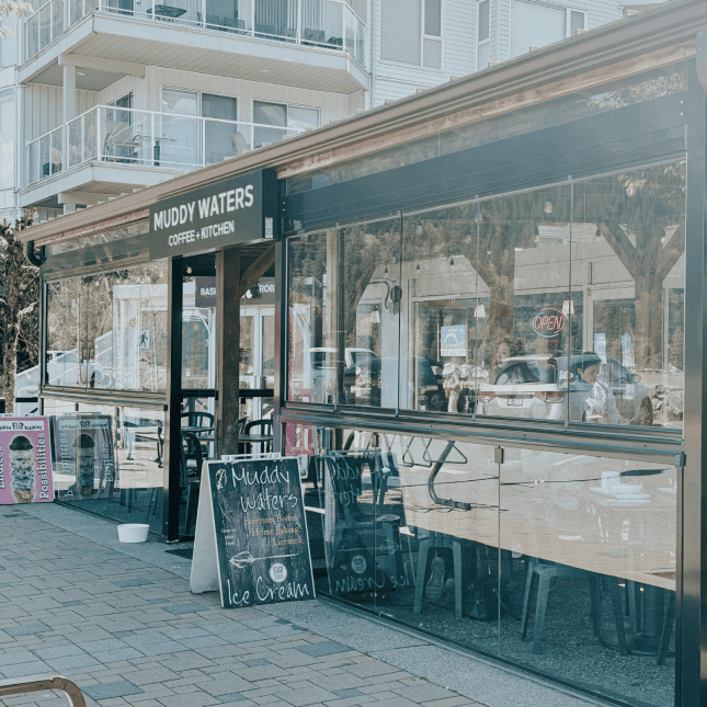 Exterior of a café storefront with large windows, outdoor seating, and signage.
