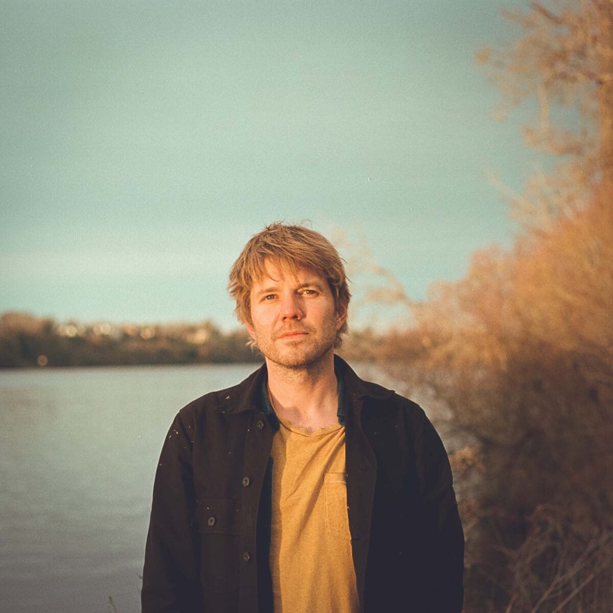 a man looking at the camera, standing in front of a body of water