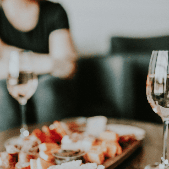 A wooden charcuterie board with cheese, fruit, and crackers on a table, with two glasses of white wine in the foreground